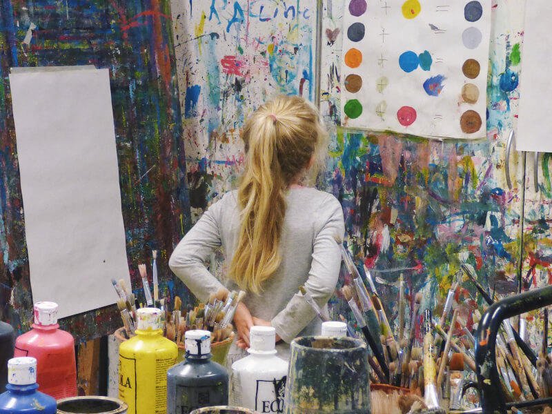 Woman staring at paint covered wall next to blank canvas and table full of paints